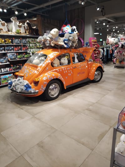 An orange Volkswagen Beetle, adorned with numerous stuffed animals, is displayed on a tiled floor within a store. The car's hood is open, revealing more stuffed animals. The stuffed animals are strategically placed on the car's roof, in the windows, and on the rear bumper. The store appears to be a toy or children's clothing store, with shelves of merchandise visible in the background, including what appear to be toy sets and clothing racks. The lighting is artificial, indicative of an indoor retail environment. The store is located in Cuxhaven, Germany.