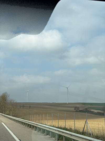 The scene depicts a rural landscape near Pagny-sur-Meuse, France, viewed from inside a vehicle. The upper left quadrant shows a section of a patterned vehicle windshield or sun visor. A paved road occupies the lower left corner, featuring a solid white line marking its edge. To the right of the road, a silver metal guardrail extends horizontally across the lower mid-ground. Beyond the guardrail, a wire fence with vertical metal posts encloses a field. The mid-ground consists of expansive agricultural fields, predominantly fallow or recently harvested with a light brown hue. Scattered deciduous trees and small clusters of vegetation are visible within these fields. In the background, two large, white, three-bladed wind turbines stand against the horizon; one is centered towards the left, and the other is partially visible on the far right. The sky is overcast with a mix of white and grey cumulus clouds against a light blue backdrop. No individuals or active subjects are present in the frame.
