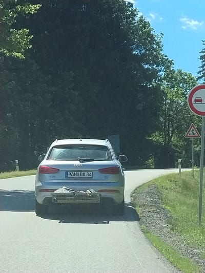 A silver Audi station wagon is parked on the side of a road, with its rear facing the viewer. The car has a license plate that reads "PAN DA 34". A metal rack is attached to the rear of the car, holding some items. The scene appears to be in a rural area, with a tree-lined road and grassy slopes. The sky is blue with some scattered clouds, suggesting it is daytime and the weather is fair. The overall atmosphere is peaceful and serene.
The image is taken from a slightly elevated angle, looking down at the car. The car is positioned at an angle to the road, as if it has just been parked or is about to be driven away. The focus is on the car and its surroundings, with the background slightly blurred. The sunlight casts shadows on the road and the car, indicating a bright day.
The presence of road signs in the background, though partially obscured, suggests that this is a public road. The trees and greenery indicate a natural environment, likely a forest or park. The road itself appears to be asphalted.
The image seems to be a snapshot taken during a road trip or a casual stop in a scenic location. There are no people visible in the image, but the car implies the presence of occupants. The overall impression is one of a pleasant drive through the countryside.
