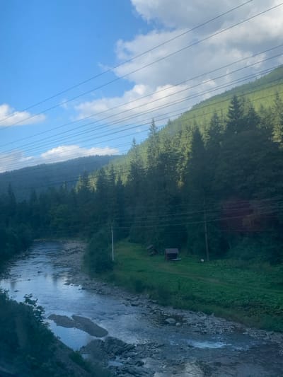 The image depicts a natural landscape in Vorokhta, Ukraine, viewed from an elevated perspective overlooking a river. The scene is dominated by a clear blue sky with scattered white cumulus clouds across the upper portion. Multiple parallel utility lines traverse horizontally across the sky and the upper half of the forested mountainside.

A rocky river, appearing to flow from the upper left to the lower right, occupies the foreground and mid-ground. Its water exhibits ripples and reflections of the sky, with numerous light-colored rocks and small gravel bars visible within its shallow bed. The riverbanks are heavily vegetated with diverse green foliage.

The right bank features a steep, densely forested mountainside covered predominantly with coniferous trees. Near the river on the right bank, there is a small, flat grassy area. Within this area, two small, dark, utilitarian structures are present: one appears to be a small, dark-roofed shed, and another less defined structure is beside it. A dirt path or cleared strip of ground is visible on the grass leading towards these structures. At least two utility poles stand on the right bank, supporting the overhead lines. The left side of the image reveals a more distant, hazy mountain range also covered in dense forest. No human figures or active interactions are visible in the scene.