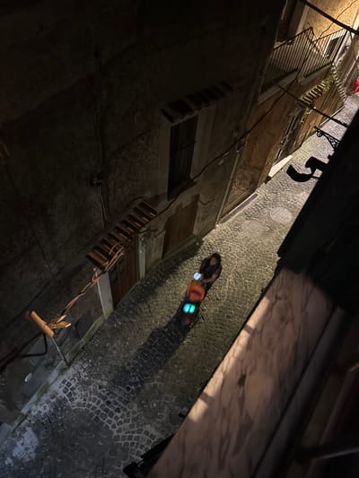 The image shows a view of a narrow street in Castelnuovo di Porto, Italy, at night. The cobblestone street is lit dimly, reflecting light from an unidentified source. Pedestrians are walking on the street. Buildings line the street, and a ladder is visible extending from one of the buildings. There are objects in the sky, but they are obscured.