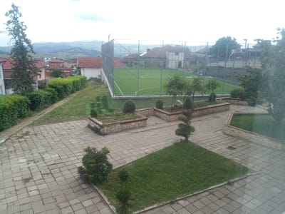The image captures a view of a schoolyard or recreational area in Valkosel, Bulgaria. In the background, a fenced, artificial turf soccer field with white markings is visible, bordered by a metal fence and situated behind residential buildings with red-tiled roofs. A tall evergreen tree stands to the left, and various hedges and manicured bushes are arranged throughout the foreground and mid-ground. The ground in the foreground is paved with square, grey concrete tiles, some of which appear uneven and worn. Several elevated, rectangular garden beds with stone borders are present, containing soil and some sparse vegetation. A section of green grass is also visible in the lower right portion of the image. The overall impression is of an outdoor space designed for sports and leisure, with a mix of paved and green areas. The sky is overcast, suggesting a cloudy day.