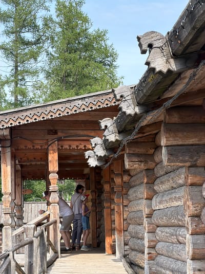 The image shows a wooden structure with intricate carvings, possibly a gazebo or a part of a traditional Russian house, located in Nevskii Parkleskhoz, Russia. Several people are present, appearing to be tourists observing the architecture. The building features logs stacked horizontally, and decorative wooden elements adorn the roof and porch. The surrounding environment includes tall trees and a clear sky.