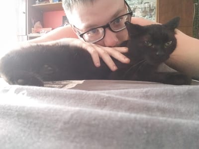 A close-up selfie shows a person wearing black-rimmed glasses and a black cat lying on a grey blanket. The person is holding the cat with their left hand. The cat is facing the camera and appears to be looking at the viewer. The background is blurred and shows a bookshelf with various items on it, suggesting the image was taken indoors. The lighting is bright, with some areas overexposed. The image is taken from a high angle, looking down at the person and cat. The specific location of Yaksha, Russia, is not identifiable from the visual information in the image.