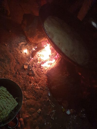 A large pot sits over a crackling fire, likely used for cooking. The fire illuminates the surrounding ground, covered in dirt and debris. In the lower right corner, a smaller pot contains what appears to be a block of dried noodles, suggesting a simple meal being prepared. The dark background suggests an outdoor cooking setting at night.