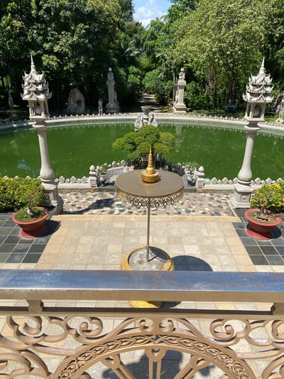 Overhead shot of a pool in Dĩ An, Vietnam, featuring green water and decorative statues along the edge. A fountain stands in the pool. Tile and potted plants decorate the surrounding area. A metal railing is in the foreground. Trees are visible in the background.