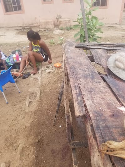 The scene is an outdoor area in Pati Ridge, Nigeria, located at coordinates 7.81 latitude and 6.704 longitude. A young woman in a black and yellow tank top and black shorts is sitting on a concrete block, holding a stick. Another person is partially visible in the bottom left corner of the frame, wearing a dark shirt. A blue plastic chair is in the foreground, along with a small orange plate and several other objects, including a bottle of water and a plastic bag, scattered on the ground. A wooden structure, possibly a table, is positioned to the right of the woman, partially obscured by a large tree in the background. The ground is dirt.