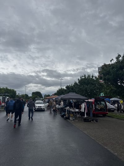 The image captures an outdoor flea market or street market scene in Groß-Gerau, Germany, under an overcast sky. The perspective is down a paved street where market stalls are set up along the right side, flanked by green trees.

Numerous people are visible, some walking on the asphalt road, and others browsing the goods at the stalls. In the foreground, a group of three individuals walks away from the camera. Further along the street, another person in a grey top walks towards a white car parked among the stalls.

The market stalls consist of various tents and display racks laden with items, including clothing. Several cars are integrated into the market setup, with vendors seemingly selling goods from their trunks. A red car with its trunk open is prominent near the middle-right. A white car, further down, also has its trunk open, and its German license plate "GG-M 3266" is clearly visible, confirming the Groß-Gerau location.

The weather is characterized by heavy, grey clouds covering the entire sky, suggesting an overcast day; the asphalt road appears damp. Lush green trees line the market area behind the stalls, and tall streetlights punctuate the scene. A large, partially obscured sign featuring a prominent "D" and possibly "DEGO" can be glimpsed through the trees.