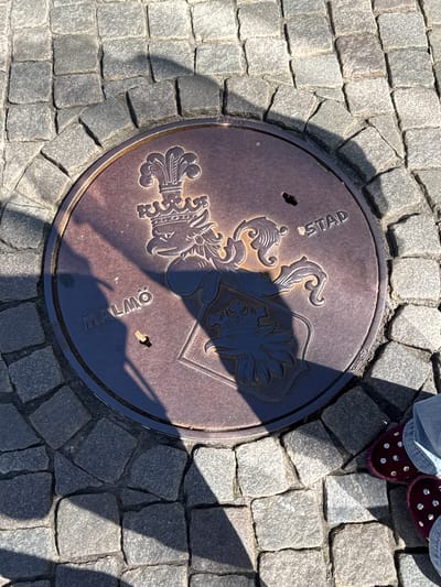 A top-down shot shows a circular metal manhole cover embedded in a cobblestone street. The cover features a coat of arms design and the text "STAD" and "MALMO." Part of a person's legs and feet, wearing jeans and maroon, studded shoes, are visible in the upper right corner of the frame. The cobblestones surrounding the cover are gray and irregularly shaped. Sunlight casts shadows across the scene. This is likely in Malmö, Sweden, based on the "MALMO" inscription.