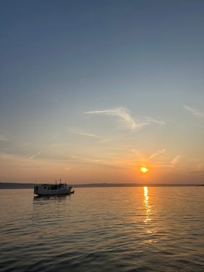 The image depicts a serene sunset over a body of water, likely a large river or lake, with a small white boat anchored on the left side. The sun is low on the horizon, casting a warm, golden glow across the scene and reflecting a shimmering path on the water's surface. The sky transitions from a clear blue at the top to soft oranges and yellows near the horizon, with wispy clouds adding texture. A faint, dark line of trees or land is visible in the distance along the horizon. There are no people visible in the image. The overall impression is one of tranquility and natural beauty. The location is the city of Sylva, Russia.