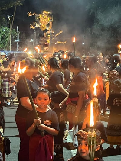 The image depicts an outdoor night scene in Munggu, Indonesia, during what appears to be a ceremonial procession or festival. A dense crowd of individuals, including adults and children, occupies the foreground and midground. Prominently featured in the center-left foreground is a young girl, wearing a black t-shirt with a white logo and a red sarong-like lower garment, holding a tall, lit bamboo torch. She directly faces the viewer. Partially visible in the lower right, another child, also holding a lit bamboo torch and wearing a patterned headband, looks forward. Most adults and children present wear dark-colored clothing. Many individuals are seen holding single or multiple bamboo poles, several of which are ignited as torches, providing the primary illumination. In the background, a large, multi-tiered effigy, consistent with an Ogoh-ogoh, stands prominently. This effigy portrays a humanoid figure with a yellow/gold face, sharp teeth, and outstretched clawed hands, mounted on a structural base and illuminated from below. The scene is characterized by the warm, flickering light from numerous torches, casting orange and yellow hues and generating visible smoke or haze. The background is dark, with silhouettes of trees and faint distant lights visible.