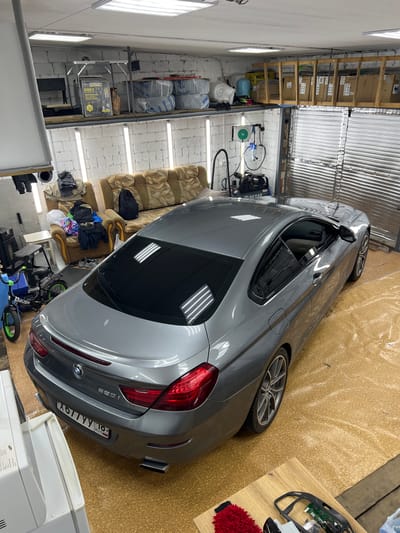 The image depicts an interior scene, possibly a garage or workshop, with a grey BMW 330i parked in the center. The car is angled, with its rear facing towards the right side of the frame. The interior is cluttered, with various items visible, including what appear to be storage shelves filled with boxes and plastic-wrapped goods, a sofa with several items of clothing draped over it, and a white appliance in the foreground. The lighting suggests an indoor environment, possibly lit by fluorescent tubes, as evidenced by the reflections on the car's surface. The floor is covered with a cork-like material.

Visible text includes "BMW" and "330i" on the car's rear. The license plate reads "X617YY 16 RUS". A label on a box on a shelf reads "KAERCHER KWD3". Another box has the brand name "HUT".

The scene appears to be a private workspace or storage area, likely in Veteran-2, Russia, given the location context. There are no people visible in the image. The overall impression is one of a functional, albeit somewhat disorganized, space used for storage and possibly car maintenance or detailing. The time of day and weather are not discernible from the image.