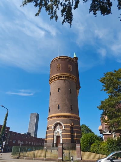 This image captures the iconic Wilhelmina Park water tower in Tilburg, Netherlands, on a clear, sunny day with a blue sky dotted with wispy clouds. The water tower, constructed from brown brick with yellow decorative bands, dominates the center of the frame. It features a cylindrical base with arched windows and doorways, tapering slightly towards the top, which is crowned by a green conical roof and a flagpole.

To the left of the water tower, a modern, tall, dark-colored skyscraper is visible in the distance, contrasting with the historic architecture. In the foreground, a street runs along the bottom, bordered by a black metal fence. A traffic light, partially obscured by foliage, stands on a pole to the left, and a row of brick buildings with numerous windows stretches horizontally across the middle ground. Lush green trees and bushes provide a natural element to the urban landscape, with some branches of a tree extending into the top of the frame from above. The scene appears to be devoid of people or any discernible action, presenting a still, architectural view of the location.
