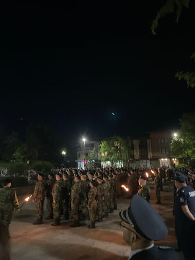 At night in Bratsigovo, Bulgaria, a large group of people in military uniforms stand in orderly rows on a paved area. Some of them hold torches. Several uniformed individuals in the foreground appear to be directing or observing the group. Buildings and streetlights are visible in the background. A crescent moon is visible in the dark sky.