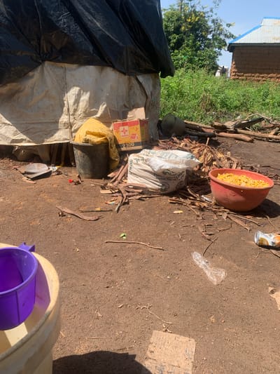 The image shows an outdoor scene, likely in Buga da Gindi, Nigeria, characterized by a dusty, dirt ground littered with debris. In the foreground, a large, light-colored bucket contains a purple scoop. To the right of this bucket, a red bowl is filled with yellow kernels, possibly corn. Further to the right and slightly behind, a discarded aluminum can lies on the ground.

Behind these foreground objects, a temporary structure is visible. It appears to be made of a large black tarp covering a lighter-colored material. To the left of this structure, a dark bucket holds a yellow bag. Near the base of the structure, a shovel and scattered pieces of wood and leaves are present. In the background, a building with a blue corrugated metal roof and mud-brick walls stands amidst lush green vegetation. The sky is visible and appears clear. There are no people clearly depicted in the scene.