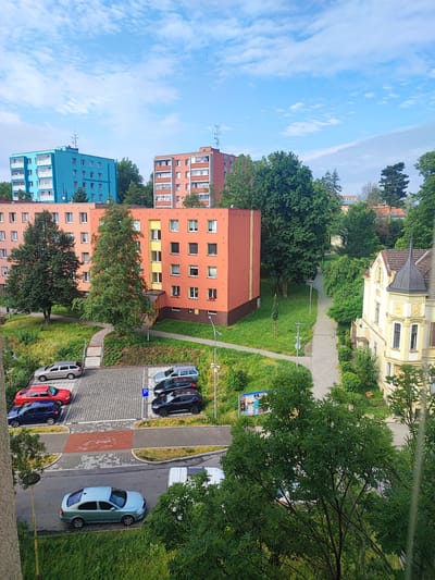 The image shows an aerial view of a residential area in Nový Jičín, Czechia. Multiple apartment buildings of varying colors, including bright teal and orange, are visible. There is a parking lot with several cars, including a red sedan, a blue hatchback, a silver station wagon, and a black SUV. A paved pathway winds through a green, grassy area with trees. A yellow building with a distinctive turret stands on the right side of the image. The sky is blue with scattered white clouds.