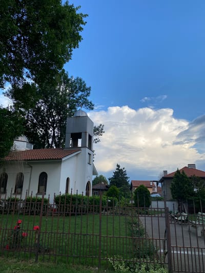 The image displays a tranquil outdoor scene in Miriantsi, Bulgaria, centered around a white church or religious building. The building features a reddish-brown tiled roof and several arched windows, some of which contain small cross motifs. A prominent bell tower, constructed from grey, possibly metallic, panels, rises from the left side of the main structure. It is topped with a dome-like roof, capped by a small cross, and has open sections for bells.

Lush green trees are abundant, framing the left side of the composition and providing a backdrop to the church. In the foreground, a dark, rusty metal fence stretches across the lower and middle sections of the image, enclosing a vibrant green lawn. Through and behind the fence, clusters of bright red roses are visible, adding a splash of color to the verdant surroundings. To the right and in the background, several residential buildings with distinctive terracotta roofs can be seen, hinting at the local architecture.

The sky is a bright blue, largely clear on the left, but dominated by large, fluffy white clouds on the right side. The lighting suggests it is daytime, possibly late afternoon, with good weather conditions. No specific activity or individuals are depicted. Faint inscriptions might be present on the church walls and bell tower, but they are too indistinct to be read. The overall impression is one of serenity and local charm.