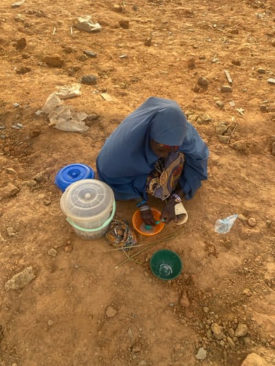 An aerial view shows a person in a blue robe and head covering washing dishes in a dusty, rocky landscape. Various containers, including a translucent bucket with a blue lid and an orange bowl, surround the individual. A pair of sandals rests near the bowl as the person's hands are submerged in the water. Debris, like plastic bags, and a green bowl are scattered nearby on the dry ground. A patterned cloth rests near the buckets.