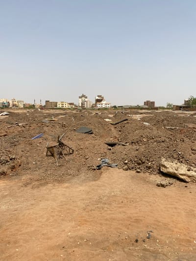 The image shows a large, flat, sandy area in Khartoum, Sudan, located at coordinates 15.528, 32.571. The area is covered in mounds of brown dirt, with a few scattered pieces of debris. In the foreground, there is a broken wheelbarrow, a blue tarp, and a piece of what appears to be a rubber tire. There are a few buildings in the background. The sky is clear and blue. There are no people present in the scene. The scene appears to be an abandoned or neglected construction site.