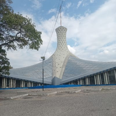 The image displays the Metropolitan Cathedral of Barquisimeto, Venezuela, under a partly cloudy sky. The main subject is the modernist cathedral, characterized by a tall, light-colored conical tower featuring a pattern of evenly spaced diamond-shaped indentations. A slender spire extends from the apex of the tower. Utility wires stretch diagonally across the sky from the left, passing near the spire. The tower integrates into a broad, multi-faceted roof structure composed of numerous light blue-grey panels. Below the roof, the building's facade is defined by vertical white and dark-colored elements, with a dark blue band visible at its base. In the immediate foreground, a cracked and weathered asphalt surface, likely a parking area, is visible, bordered by a low concrete curb painted blue. A black lamppost stands in the mid-ground, positioned to the left of the cathedral. A portion of a leafy green tree is present on the far left edge of the frame. No individuals or discernible actions are depicted.