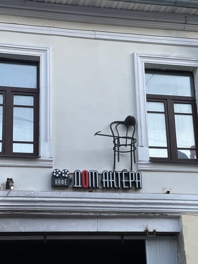 The image shows the facade of a building, likely a cafe, with two windows and a sign. The sign reads "КАФЕ ДОМ АКТЁРА" in stylized lettering, with a film reel icon to the left of "КАФЕ". Above the sign, an artistic metal sculpture of a chair with a hat on it is mounted on the wall. The building is painted in a light, neutral color. The scene appears to be outdoors, with overhead wires visible near the windows. No people are present in the image. The location is identified as Yazvitsevo, Russia.