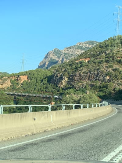 The image depicts a scenic view of a mountain landscape and a highway in Esparreguera, Spain. The foreground features a concrete highway barrier with a light blue railing, guiding the flow of traffic around a curved section of road. The asphalt surface of the road is visible, marked with white lines.
Beyond the highway, a steep, rocky mountain face is covered in dense green vegetation. A prominent electrical tower with high-voltage wires stretches across the sky, showcasing the infrastructure present in the area. The mountain exhibits rugged textures and varying shades of green and brown.
Further into the distance, more mountainous terrain is visible, with some peaks appearing sharp and defined against a clear blue sky. The presence of the electrical tower and the road suggests a natural landscape that has been adapted for human use, possibly a scenic route or a passage through the mountains.
The lighting suggests it is daytime, likely midday or afternoon, with the sun casting clear shadows and illuminating the scene brightly. The weather appears to be clear and sunny, conducive to outdoor activities and travel. There are no people visible in the image, but the presence of the road implies ongoing vehicular activity. The overall impression is one of a beautiful, natural environment integrated with man-made structures.