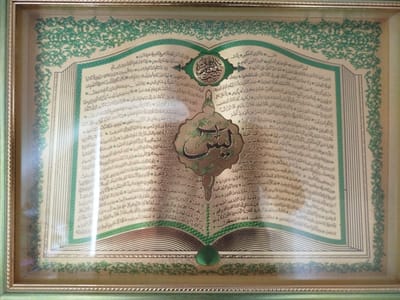 This is a framed piece of Islamic calligraphy, likely a framed Quranic verse or devotional text. The artwork is presented as an open book, with text in Arabic script filling the pages. The calligraphy itself is elaborate and decorative, with gold lettering and intricate green floral motifs framing the text and adorning the central elements. A prominent, oval-shaped emblem with swirling calligraphy is positioned near the top center of the "open book," and a similar, though larger, decorative element appears below it. The entire piece is set within a light green and gold frame with a textured border. The artwork is displayed behind glass, which reflects the light and the surrounding environment, showing hints of a room. The location context provided is Ziyanchurino, Russia, suggesting this is a decorative item found in a home or religious space there. The overall impression is one of religious devotion and artistic craftsmanship.