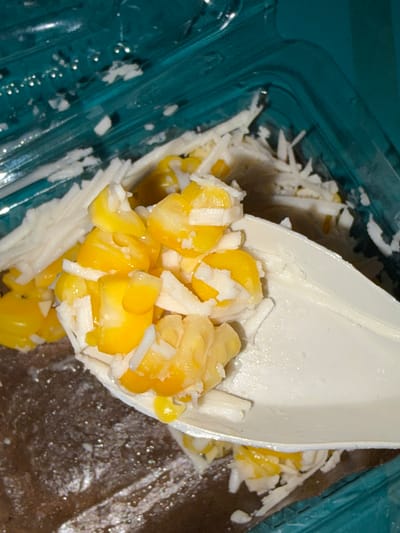 A close-up shot shows a white plastic spoon filled with corn and shredded white substance, possibly cheese or coconut, against a dark brown textured background. The spoon is positioned over a clear plastic container with a teal-colored lid and sides. The container appears to hold more of the corn and shredded white substance. The lighting is direct and harsh, creating bright highlights on the food and spoon, and casting shadows within the container. The scene is indoors, with no view of the external environment or city of Tupah, Indonesia.