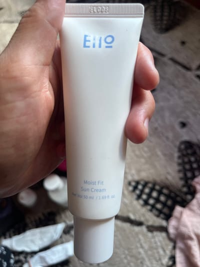 A close-up shot shows a person's hand holding a white tube of "Moist Fit Sun Cream." The tube is oriented horizontally, with the cap on the right. The brand name "ΕΙΠΩ" is prominently displayed in blue on the left side of the tube, with "ELOOS" printed vertically to its left. Text to the right of the brand name reads "Moist Fit Sun Cream" and "Net Vol 50 ml / 1.69 fl. oz." The background is blurred, showing a patterned fabric, possibly a rug or blanket, with a pink cloth draped over it. The lighting suggests an indoor setting, possibly during the daytime. No specific location cues are visible beyond the provided context of Tashlak, Kyrgyzstan. The image focuses solely on the product and the hand holding it, giving no indication of any activity or event. The overall impression is a straightforward product shot.