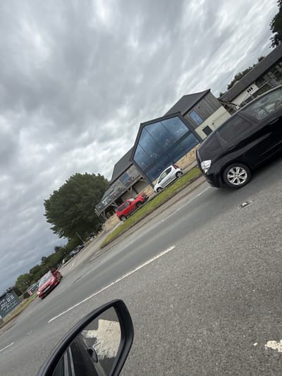 This media file presents an outdoor scene along a road in Long Ashton, United Kingdom, under an overcast sky. The image is captured from inside a vehicle, with its black wing mirror prominently displayed in the lower right corner, reflecting a white road marking and the asphalt surface.

The main subject is a modern building situated on the left side of the road. This establishment features extensive glass panels, a dark roof, and dark-toned cladding, giving it a contemporary architectural style. A sign in the lower right foreground, partially obscured by the wing mirror, identifies the building as "THE INN at [partially visible] PUB & ROOMS."

Several cars are parked along the road and in front of the inn. In the left foreground, a black hatchback or SUV is parked partially on a grass verge. Further down the road, a white car and a red SUV (resembling a Jeep Wrangler) are visible. The road itself is made of asphalt with distinct white line markings.

Lush green trees and grass verges line the road and are present around the building, suggesting a rural or suburban setting. The sky is predominantly grey and cloudy, indicating an overcast day. No individuals are explicitly visible in the frame, but the parked cars imply human activity, likely guests or patrons of the inn.
