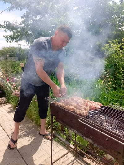 A man is shown barbecuing meat on skewers over a grill in a backyard in Vyazovka, Russia. He is wearing a dark t-shirt and dark pants, with flip-flops on his feet. The man has tattoos on his left arm and is looking down at the meat as he adjusts it with tongs.

The scene is set outdoors, with lush green foliage and flowers visible in the background. Smoke rises from the grill, obscuring some of the background and creating a hazy atmosphere. The ground is paved with light-colored bricks. The lighting suggests it is daytime, likely sunny. The overall mood appears casual and relaxed, indicative of a leisure activity. There is no visible text in the image.