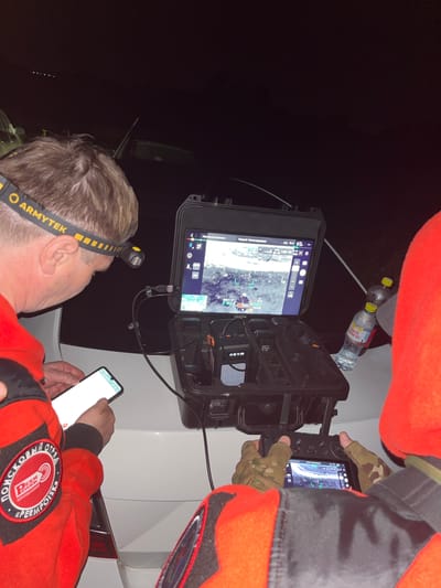 In Badashka, Russia, a team is engaged in a nighttime search operation. A man wearing a yellow and black Armytek headlamp, a red jacket with a "POISKOVY OTRYAD PERM POISK" patch, is looking at a smartphone. Another person, also in a red jacket, is operating a drone controller with a screen displaying a thermal image. The drone controller and a ruggedized case containing drone equipment are placed on the trunk of a white car. Two water bottles, one labeled "SAINT SPRING," are also visible. The scene is illuminated by the headlamp and the screens, suggesting a focus on operations in low-light conditions.
