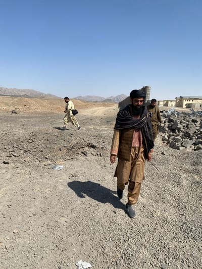 The image captures an outdoor scene in Bar Mel, Afghanistan, under a clear, cloudless blue sky. Three adult males are walking across an uneven, rocky, light grey-brown ground surface. In the foreground, a man with a dark beard, a dark turban, and a dark patterned shawl draped over a patterned vest and light brown tunic and trousers, walks towards the lower right, holding a thin dark stick in his right hand. He wears dark slip-on shoes, and his shadow is cast strongly to his left. Behind him and to his right, a second man with a beard, wearing a khaki-colored tunic and trousers, follows, walking in a similar direction. To the mid-left, a third man with a beard, dressed in a light-colored tunic and trousers, walks from left to right with a dark shoulder bag slung across his body. In the mid-ground on the right, a large pile of broken concrete blocks is visible, alongside a partially damaged concrete wall structure. Further in the background on the right, several light-colored, multi-story buildings, appearing either under construction or damaged, are present. Distant arid mountains form the horizon under direct sunlight, which creates crisp shadows.