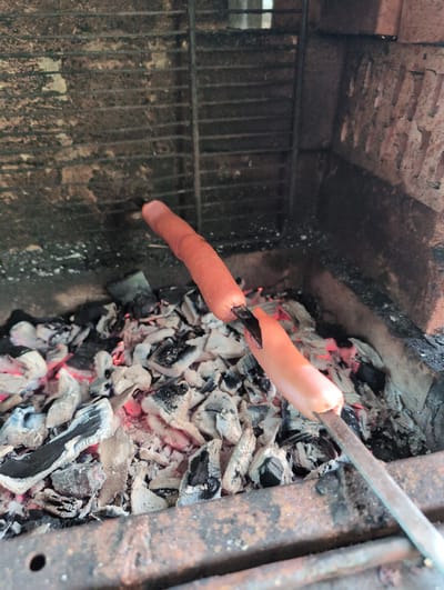 In the image, a person is grilling hot dogs over hot coals. The setting appears to be an outdoor grill or fire pit with a brick background and a metal grate. The hot dogs are impaled on a metal skewer, and one is being held close to the coals, sizzling. The coals are glowing red and emitting heat, suggesting the grilling is in progress. The lighting is natural, indicating it's likely daytime. The image captures a moment of cooking and outdoor activity, possibly a casual gathering or a meal preparation. There is no visible text in the image. The location context provided is Vyishki, Russia.