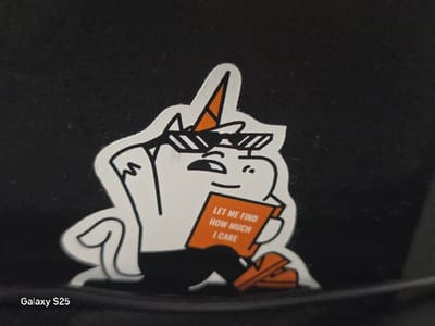 The image displays a white cartoon sticker featuring a unicorn character, seated, against a solid black background. The character has black outlines, an orange horn with black stripes, and wears black sunglasses with white diagonal lines, black pants, and orange shoes. It holds an open orange book with white text that reads "LET ME FIND HOW MUCH I CARE". The sticker has a distinct white border. In the bottom-left corner of the image, a "Galaxy S25" watermark is visible. The scene is located in Novo Delchevo, Bulgaria.