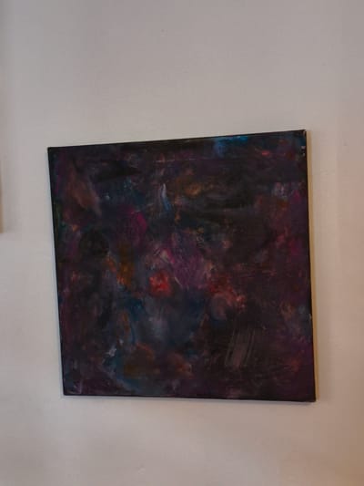 A square, abstract painting with dark, swirling colors of purple, blue, pink, and hints of red and orange is mounted on a plain, lightly textured off-white wall. The painting has a rough, impasto texture with visible brushstrokes. The image does not contain any information that allows for determination of its location in Cavite City, Philippines.