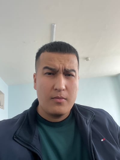 A man faces the camera with a furrowed brow and a slightly skeptical expression. He has a very short, close-cropped haircut and appears to be wearing a dark blue zip-up jacket over a green crewneck sweater. A small red and white logo is visible on the jacket. The background consists of a pale, off-white wall with some visible fixtures. The image is oriented at a slight angle.