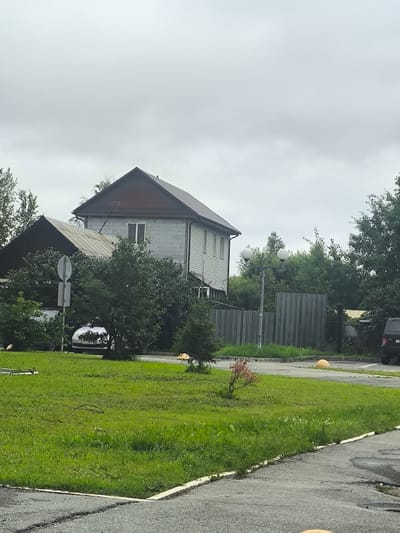 The image depicts a cloudy day in Sredneuralsk, Russia, likely on the outskirts of the city, near a residential area. In the background, a two-story house is visible, constructed with light-colored cinder blocks and featuring a dark brown, sloped roof. The house has at least one window on the upper floor. Adjacent to this house is another structure with a similarly dark, sloped roof, partially obscured by lush green trees and bushes. A gray metal fence runs along the property line, separating the yard from a paved area.
In the foreground, a well-maintained green lawn occupies the majority of the space. A small, reddish-leafed bush is planted on the lawn. A road or parking lot with cracked asphalt is present in the bottom portion of the image. Several large, yellow, spherical bollards are visible on the paved areas. A silver car is parked behind some bushes, and the rear of a dark-colored SUV is parked on the right side of the image. A white circular sign on a pole is also visible amidst the greenery. The overall impression is that of a quiet, possibly suburban or semi-rural, setting under an overcast sky.