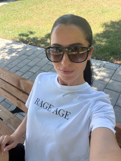 A woman with dark hair pulled back and wearing large sunglasses and a white t-shirt with the words "RAGE AGE" printed on it sits on a wooden bench in a park. The bench is made of light-colored wood and has visible screws. Behind her, a paved walkway made of grey rectangular stones is visible, and beyond that, a grassy area with dappled sunlight. The overall setting suggests an outdoor urban environment, potentially a public park in Chavdartsi, Bulgaria, during daytime.