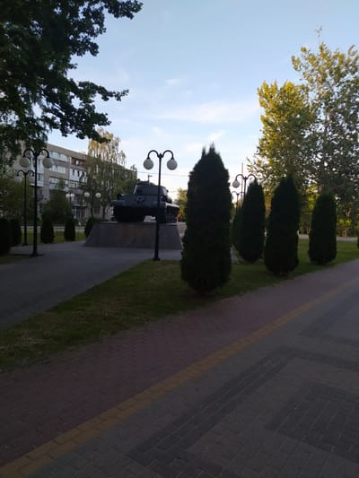 This is a photograph of a monument featuring a tank, likely in Gusev, Russia. The monument is situated on a paved walkway with a grassy area and a line of trees. The tank appears to be a military exhibit, standing on a stone pedestal. Multiple lampposts with spherical lights line the walkway, suggesting an outdoor public space. In the background, a multi-story building with numerous windows is visible, indicating an urban or suburban setting. The sky is clear and blue with a few wispy clouds, and the sunlight suggests it is either morning or late afternoon. The overall scene evokes a sense of solemnity and historical remembrance, fitting for a military monument. There is no visible text in the image.