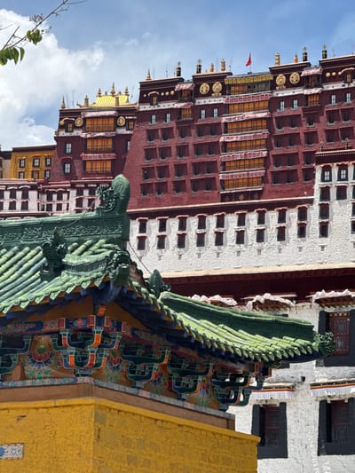 The image captures a detailed view of a grand architectural complex, strongly resembling the Potala Palace in Lhasa, Chengguan District, China, under a bright blue sky with sparse white clouds. The main structure features a striking contrast between its deep red and white sections.
The predominantly red section, visible on the left and in the background, is characterized by numerous rectangular windows, many highlighted with golden frames, and topped with ornate golden roof ornaments. A small red flag can be seen flying from one of its upper terraces. Adjacent to this, a white building section, textured and with smaller, dark-framed windows, stretches across the middle ground.
In the foreground, an elaborate green-tiled roof takes prominence on the right. This roof displays intricate patterns of ridged green tiles and features ornate decorative figures, possibly mythical creatures or gargoyles, along its ridge and at its corners. The eaves beneath the green roof are adorned with vibrant, multi-colored paintings showcasing traditional patterns in blue, red, green, and yellow. To the far right, a textured yellow wall or building facade is partially visible. A few leafy branches extend into the bottom-left corner of the frame, hinting at nearby vegetation. The bright, clear lighting indicates a daytime shot, likely midday or early afternoon. There are no visible people, activities, or text in the image.