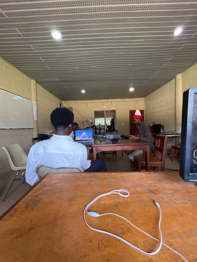 The image depicts the interior of a room, likely a classroom or computer lab, in Gembu, Nigeria. In the foreground, a wooden table is visible, with a pair of white earbuds resting on its surface. A person wearing a white shirt is seated at a desk in the middle ground, with their back to the camera, facing a laptop that is open and displaying a login screen. To the right of this individual, another person is seated at a desk and appears to be typing on a keyboard. This second person is wearing a red and white hat. The room has a suspended ceiling with fluorescent lighting and walls constructed of painted cinder blocks. A whiteboard is mounted on the wall in the background to the left. The overall impression is of an educational or training environment.