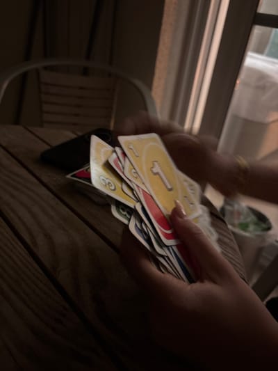 This image depicts a close-up, dimly lit scene of a card game in progress on a wooden slatted table, situated in Selçuk, Turkey. A person's left hand, featuring light-colored nail polish, holds a fanned array of Uno cards, prominently displaying a yellow '1' and a yellow '3' card, alongside red and other obscured cards. A dark-colored smartphone lies horizontally on the table to the left of the cards. In the upper right, a blurred arm adorned with a yellow bracelet is partially visible, suggesting the presence of a second individual. The background reveals a light-colored metal-framed chair with wooden slats and a window or glass door frame, providing ambient light. A white container or bucket with a green insert is vaguely visible in the far right background.