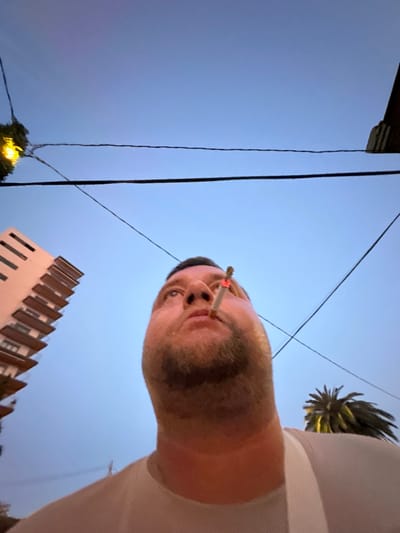 In this low-angle, close-up shot taken in Sant Feliu De Guíxols, Spain, a man is captured looking upwards with a lit cigarette in his mouth. The background reveals a clear blue sky, bisected by several dark power lines. To the left, a portion of a modern building with stacked balconies is visible. To the right, the distinctive fronds of a palm tree peek into the frame. The man is wearing a light-colored t-shirt.