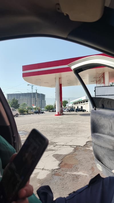 A view from inside a car shows a gas station with red and white accents. A large building under construction is visible in the background, along with a crane. Several cars are parked or driving on the asphalt lot. The sky is clear and blue. A person's hand holding a smartphone is in the foreground, obscuring part of the scene.
