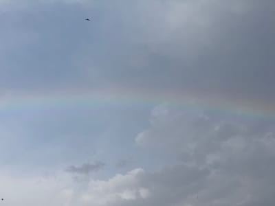 The image shows a pale rainbow in a cloudy sky. The lower portion of the sky is filled with cumulonimbus clouds, with lighter cirrus clouds above and a darker cumulonimbus formation at the bottom. A small bird is visible in flight at the top left of the frame. Another bird is barely visible in flight at the bottom left. The image was taken in Erdemli, Turkey.