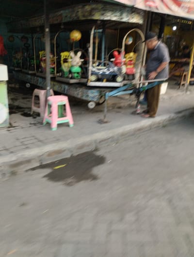 The image depicts a blurry scene in Bringinbendo, Indonesia, possibly during the daytime given the ambient light. The focus is on a small, open-air amusement ride, likely a carousel, designed for children. The ride features several colorful, cartoonish animal figures, including a rabbit, and various other whimsical characters, some of which are green. A man wearing a dark shirt and light-colored pants is visible near the ride, appearing to be interacting with it or its mechanisms. He is also wearing a hat and sandals.
In the foreground, two plastic stools are placed on a paved area. One stool is pink and green, while the other is a lighter pink or beige. The ground appears wet in patches, suggesting recent rain or a spill. The background shows more of the amusement ride structure, which is decorated with vibrant, albeit somewhat faded, paint and possibly some dangling ornaments or lights. The overall setting suggests an outdoor market, park, or a temporary entertainment setup. The image is quite blurry, making precise details of the faces or fine textures difficult to discern. No visible text is present in the image. The atmosphere seems casual, possibly indicating a local community event or a small business operating for entertainment.