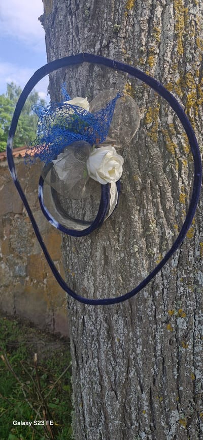 A fascinator with a blue mesh overlay and white artificial roses is attached to a tree trunk with bark in Pakruojis, Lithuania. The fascinator is secured by a dark blue velvet ribbon that encircles the trunk. The background shows a stone wall, green grass, and a bright sky with some clouds.