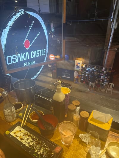 The image depicts an outdoor dining table at Osaka Castle, El Nido, Philippines. The table is covered with remnants of a meal, including dirty dishes (bowls, a plate, and small condiment dishes), chopsticks, a glass with a straw, and bottles. The sign for "Osaka Castle El Nido" is visible in the lower left. In the background, a street scene is visible with people and shops.