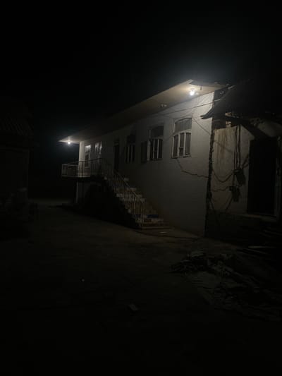 A nighttime exterior scene depicts a modest, multi-story building with a corrugated metal roof. The building is illuminated by a single light fixture above a balcony, casting a warm glow on the white facade and an outdoor staircase leading to the entrance. Several windows are visible, some with shutters. The surrounding area is dark and appears to be unpaved. To the right of the building, exposed electrical wires and conduits run along the wall, suggesting an outdoor utility box or meter. A pile of dark fabric or tarp lies on the ground in the foreground. The overall impression is of a residential structure in a rural or semi-rural setting, possibly within the village of Kedei-Aryk, Kyrgyzstan, given its aesthetic and basic infrastructure.